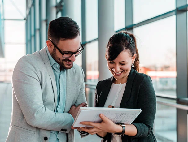 Two business professionals smiling and reviewing notes in a modern glass office hallway.